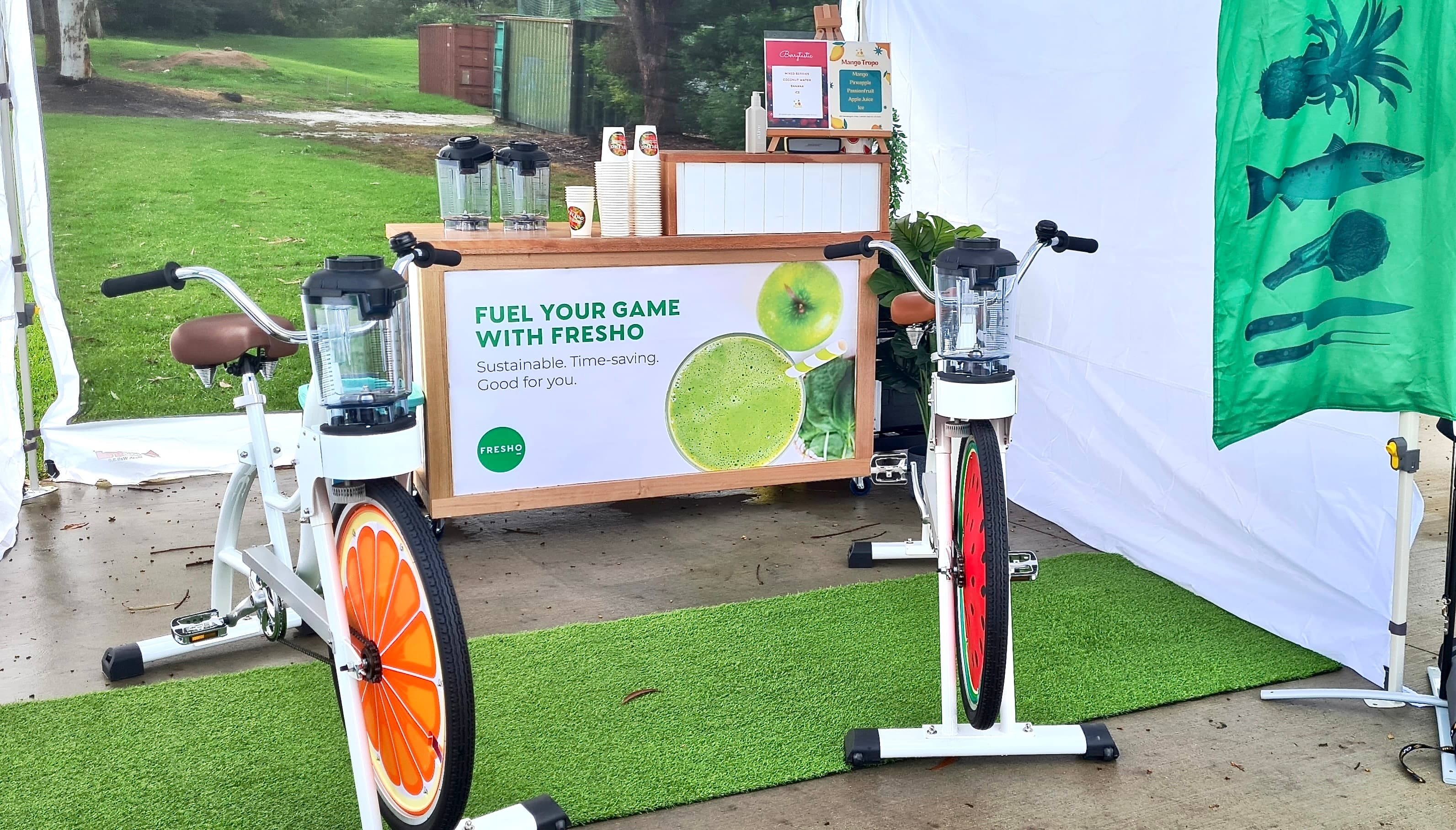 An outdoor mobile smoothie bar labeled "FUEL YOUR GAME WITH FRESHO" sits under a white tent on a rainy day. Two white stationary bikes with blender attachments are positioned in front of the bar on a strip of artificial turf. The bike wheels are decorated with vibrant orange and watermelon slice graphics.
