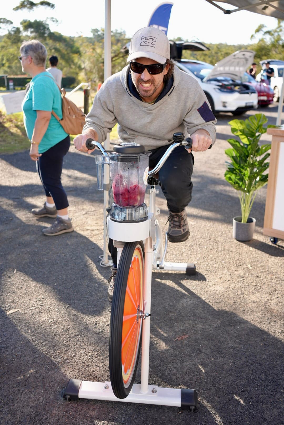 A man in a grey hoodie, sunglasses, and a baseball cap intensely pedals a smoothie bike outdoors. He is focused on blending a red berry mixture in the blender attached to his handlebars. The front wheel features an orange slice design.