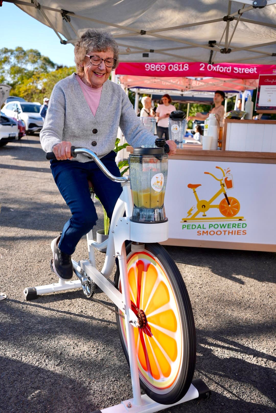 A smiling elderly person rides a stationary bike that powers a blender at a smoothie stall. The bike's wheel features a bright orange slice design.