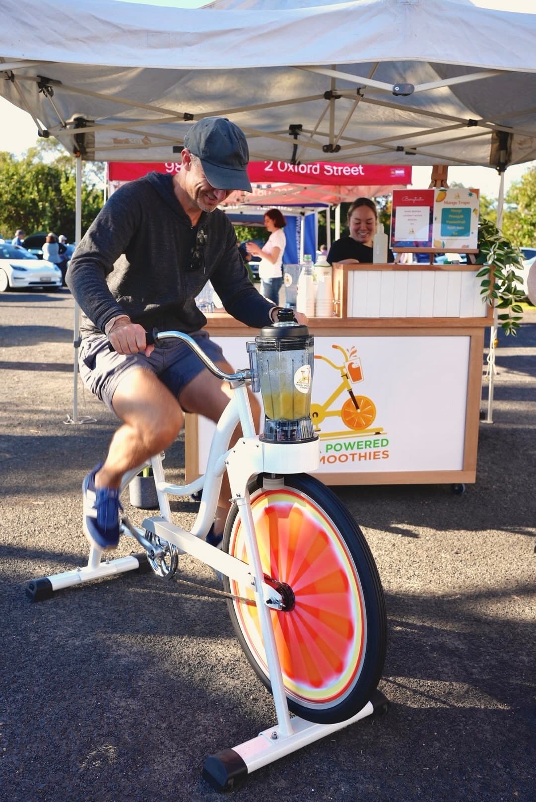 Man enthusiastically pedals a stationary bike powering a blender to make smoothies at an outdoor market stall. Vendor smiles in the background.