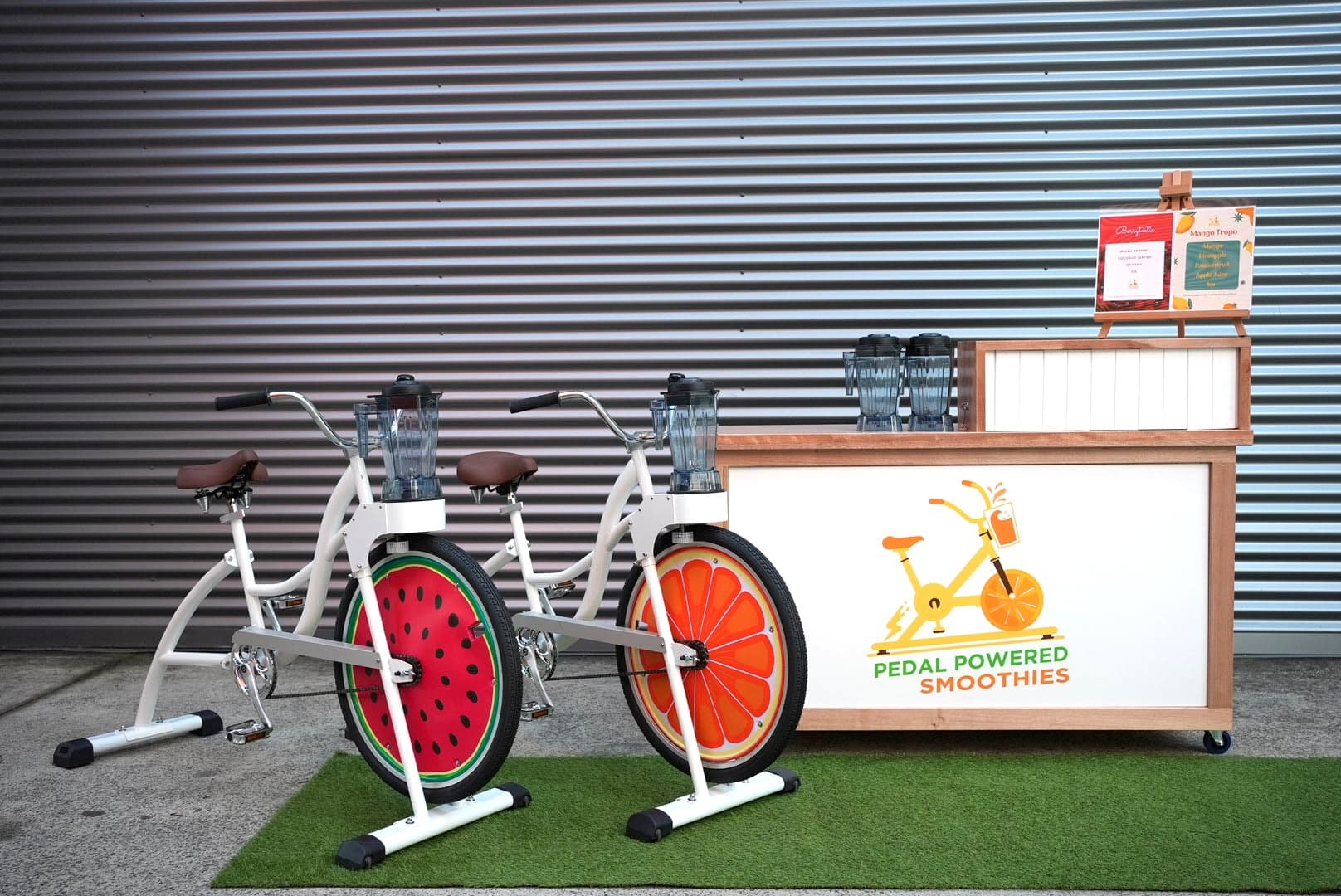Two stationary bikes with blender attachments and fruit-themed wheels are set up in front of a booth labeled “Pedal Powered Smoothies” on a patch of artificial grass.