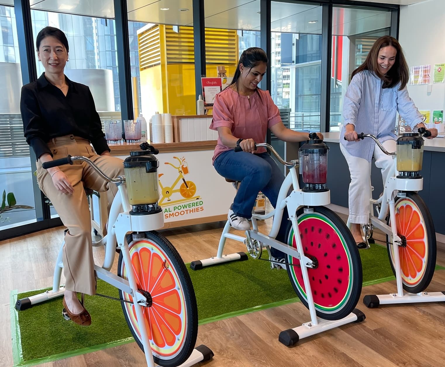 Three women on stationary bikes power blenders on a counter with "Pedal Powered Smoothies" sign in a bright, modern office space, smiling and engaged.