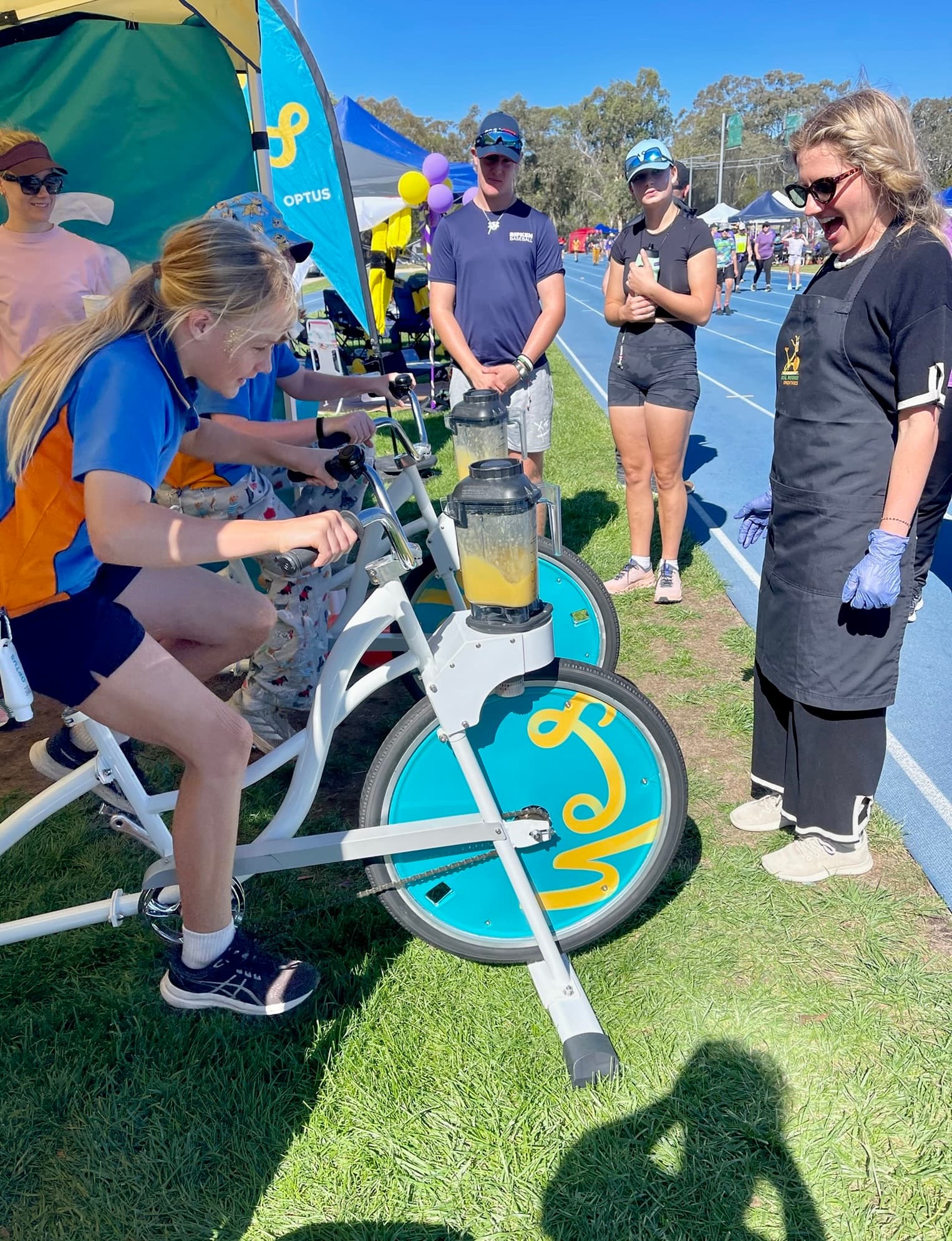 A young girl in a blue and orange athletic uniform pedals a smoothie bike at an outdoor track event. A woman in a black apron and blue gloves stands nearby, watching with an excited expression.
