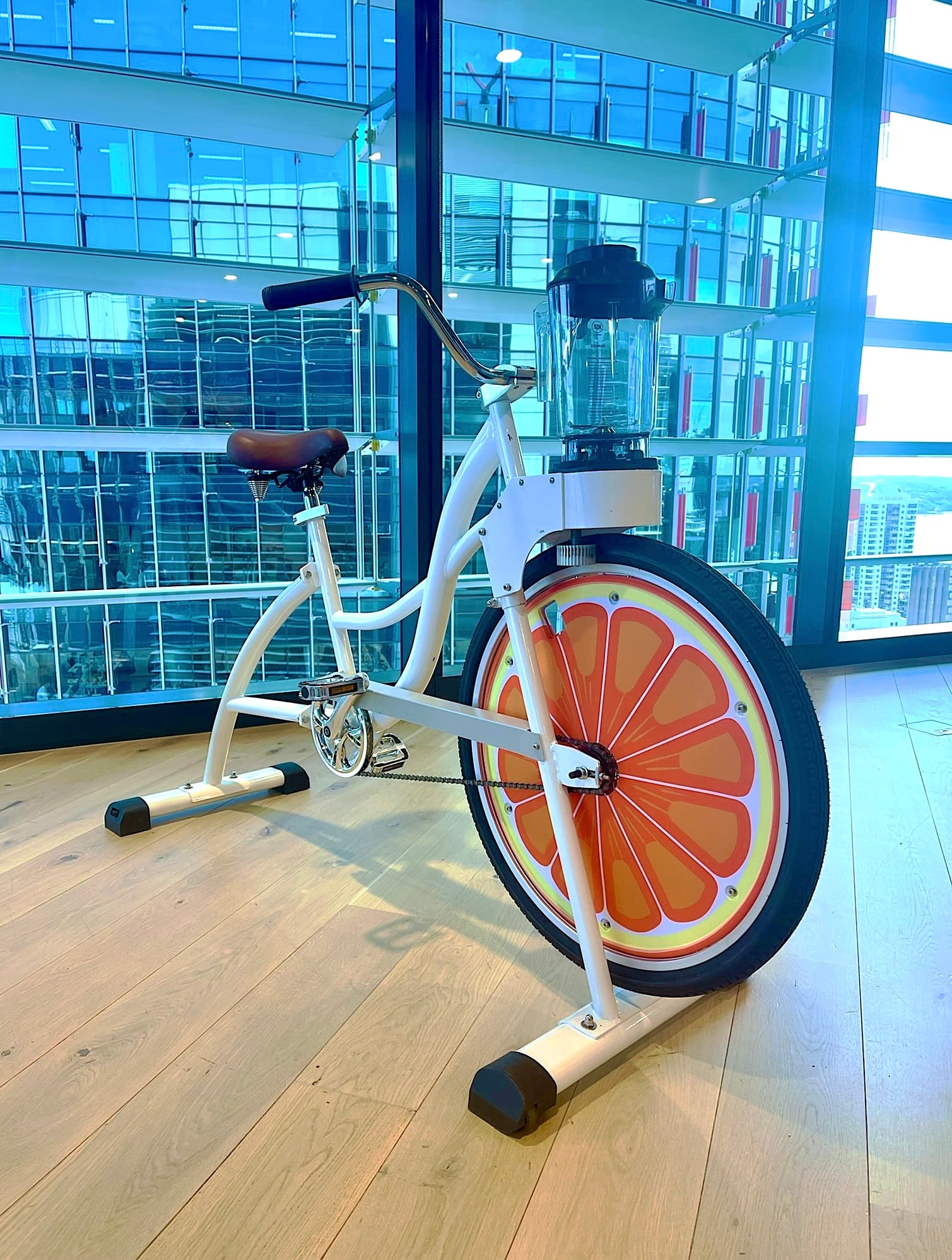 A stationary bike with a large orange slice design on the front wheel, featuring a blender attached to the handlebars, set in a modern glass-walled room.