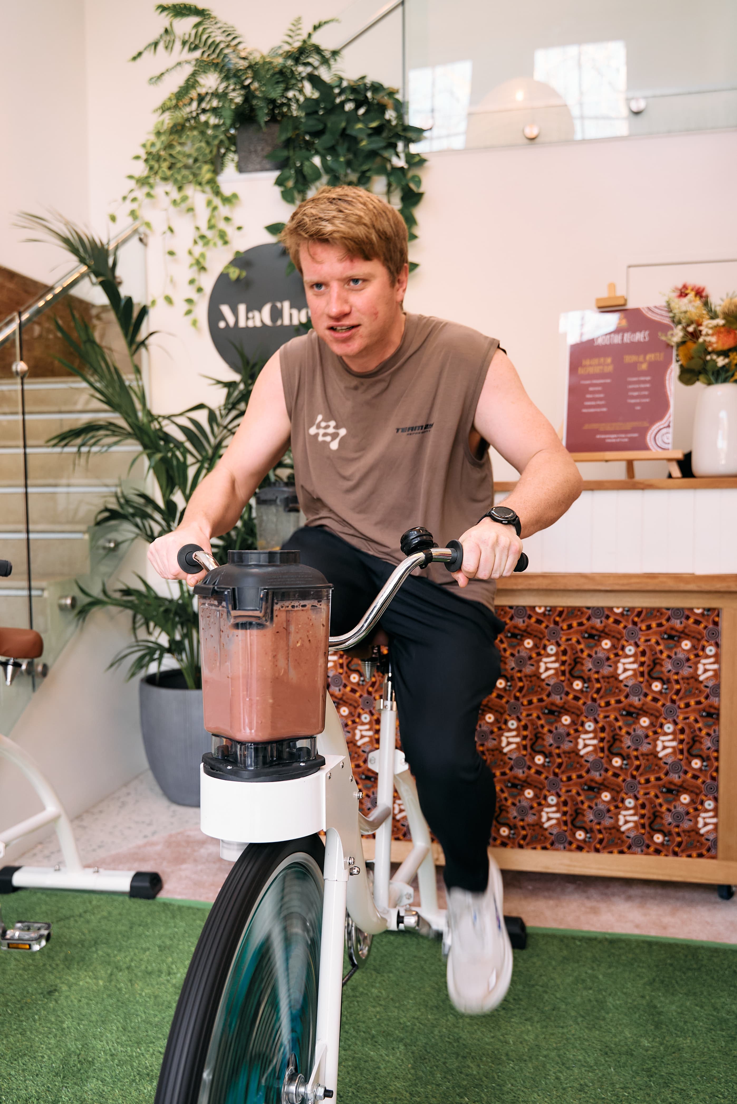 A man in a brown tank top pedals a smoothie bike indoors on a patch of artificial grass. He is looking ahead with concentration as he blends a dark brown smoothie. In the background, there is a staircase and lush green potted plants.