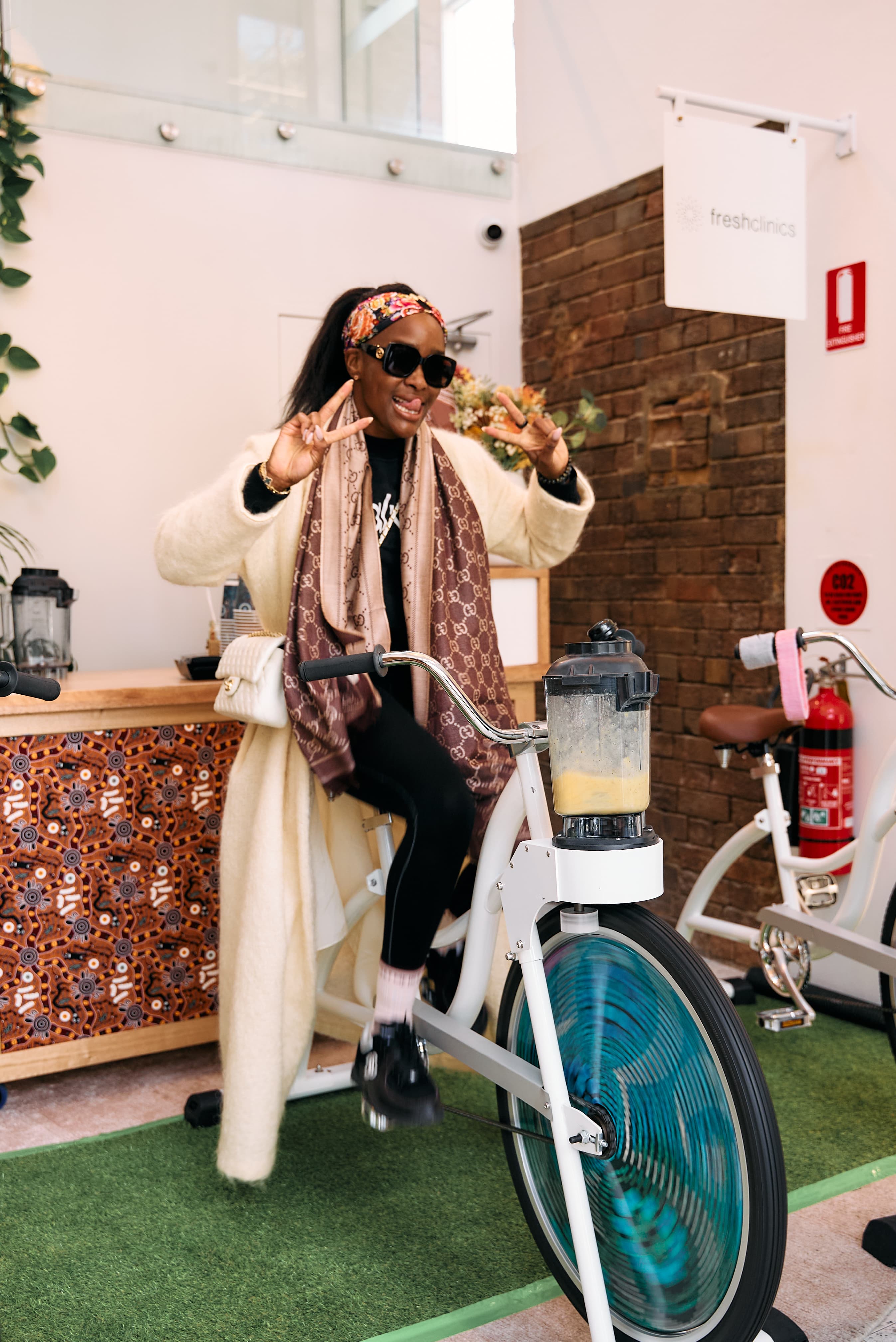 A person in a cozy outfit rides a stationary bike with a blender, making a smoothie. They're smiling and flashing peace signs indoors.