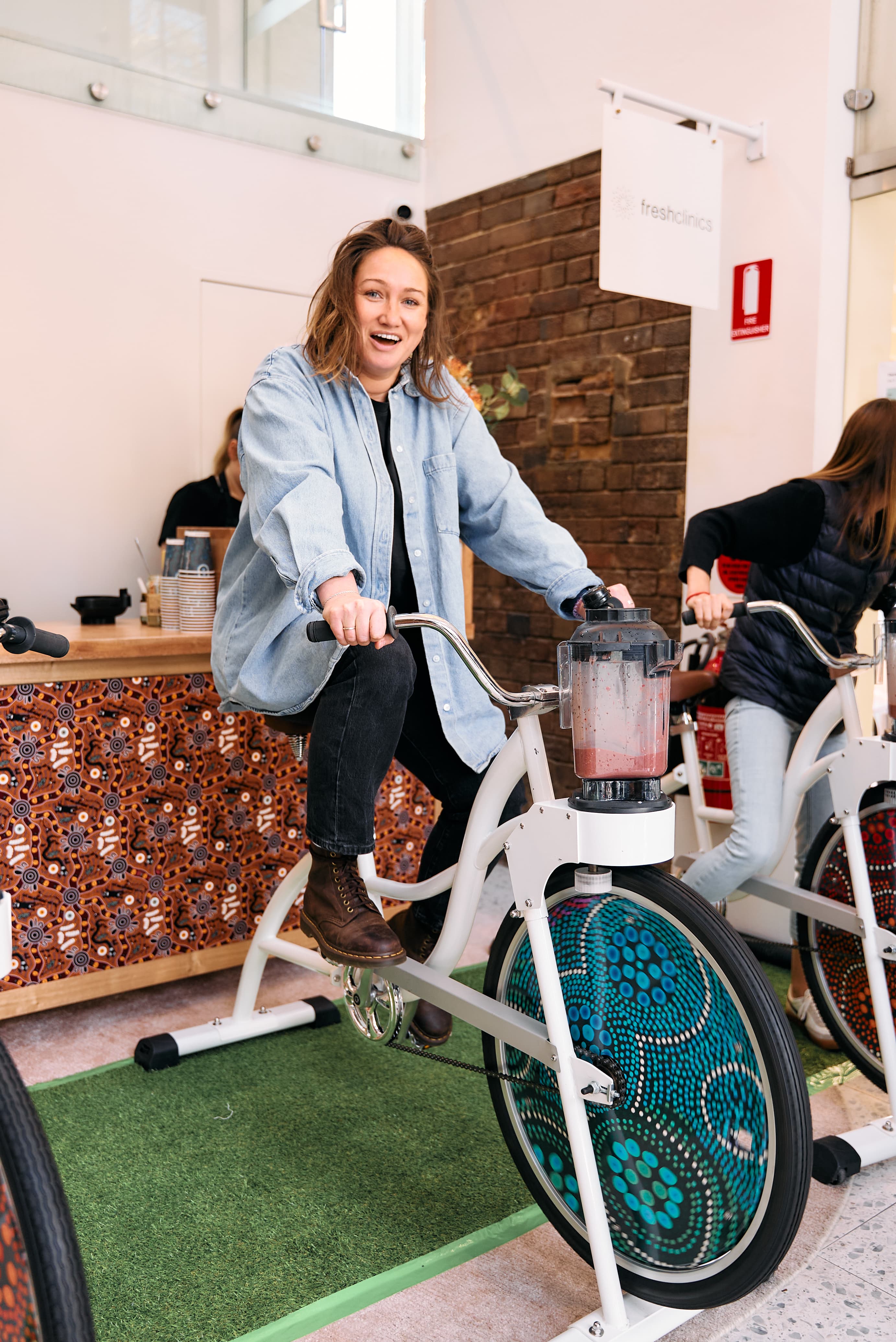 A woman in a denim shirt and black pants smiles broadly while pedalling a smoothie bike. The bike features a unique front wheel decorated with intricate Indigenous-style dot art in teal and blue.