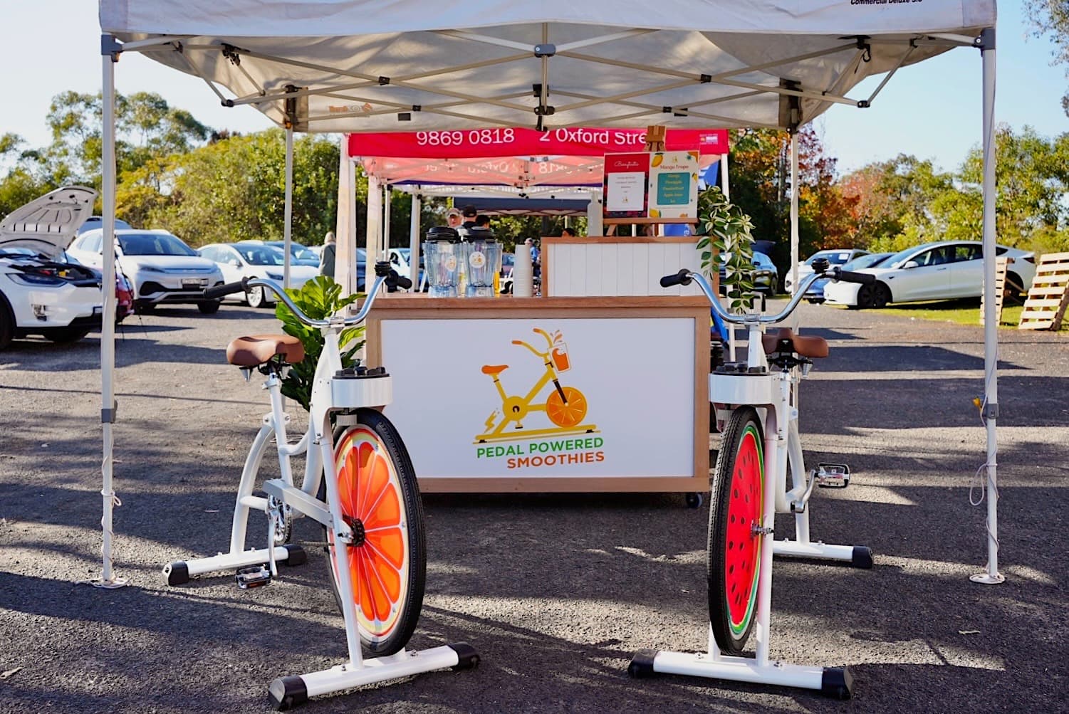 A stall with a sign that reads “Pedal Powered Smoothies” featuring stationary bikes with fruit designs on the wheels. The atmosphere is vibrant and eco-friendly.