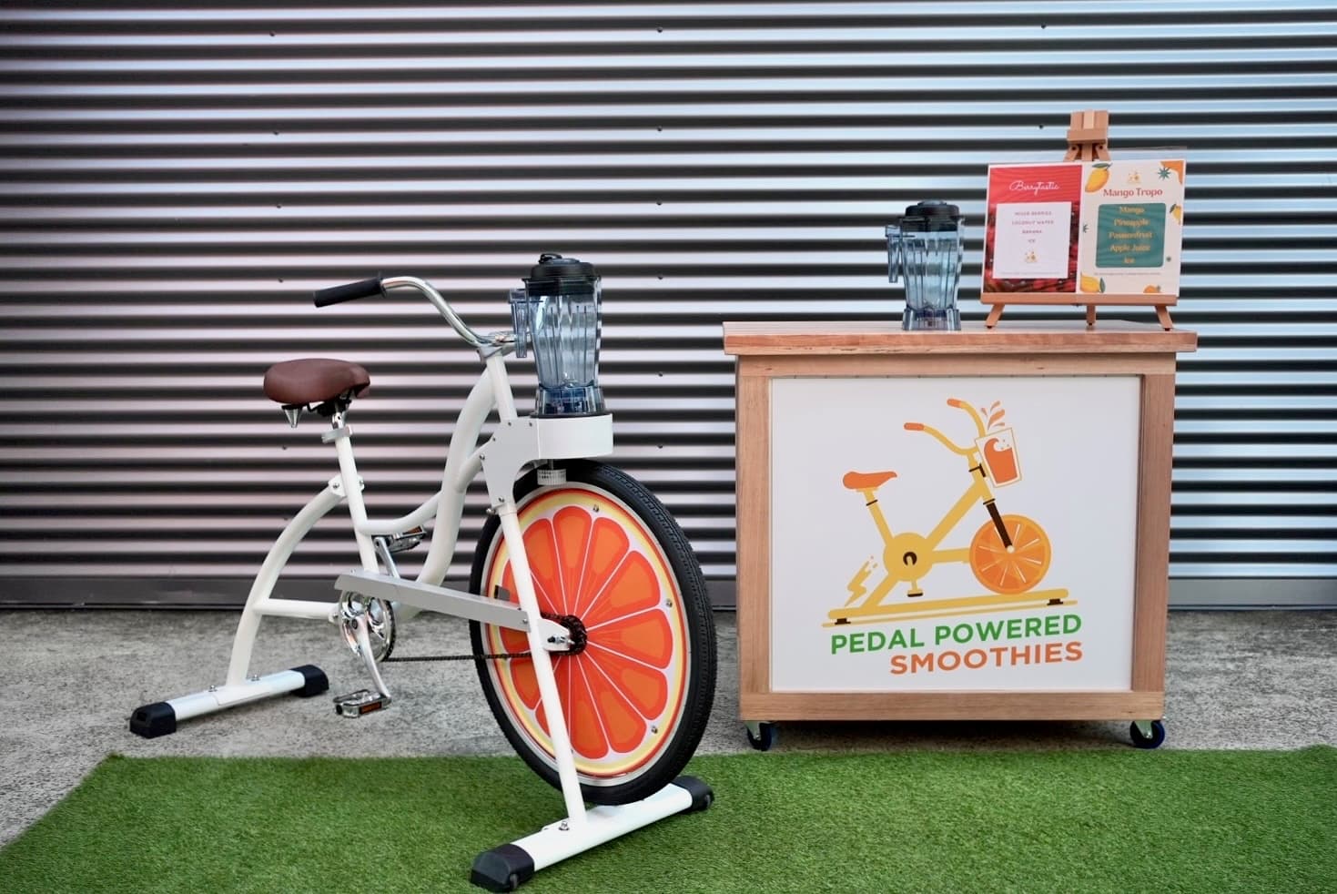 A stationary bike with a blender attached to the handlebars is next to a counter labeled “Pedal Powered Smoothies” on artificial grass, in front of a metal wall.