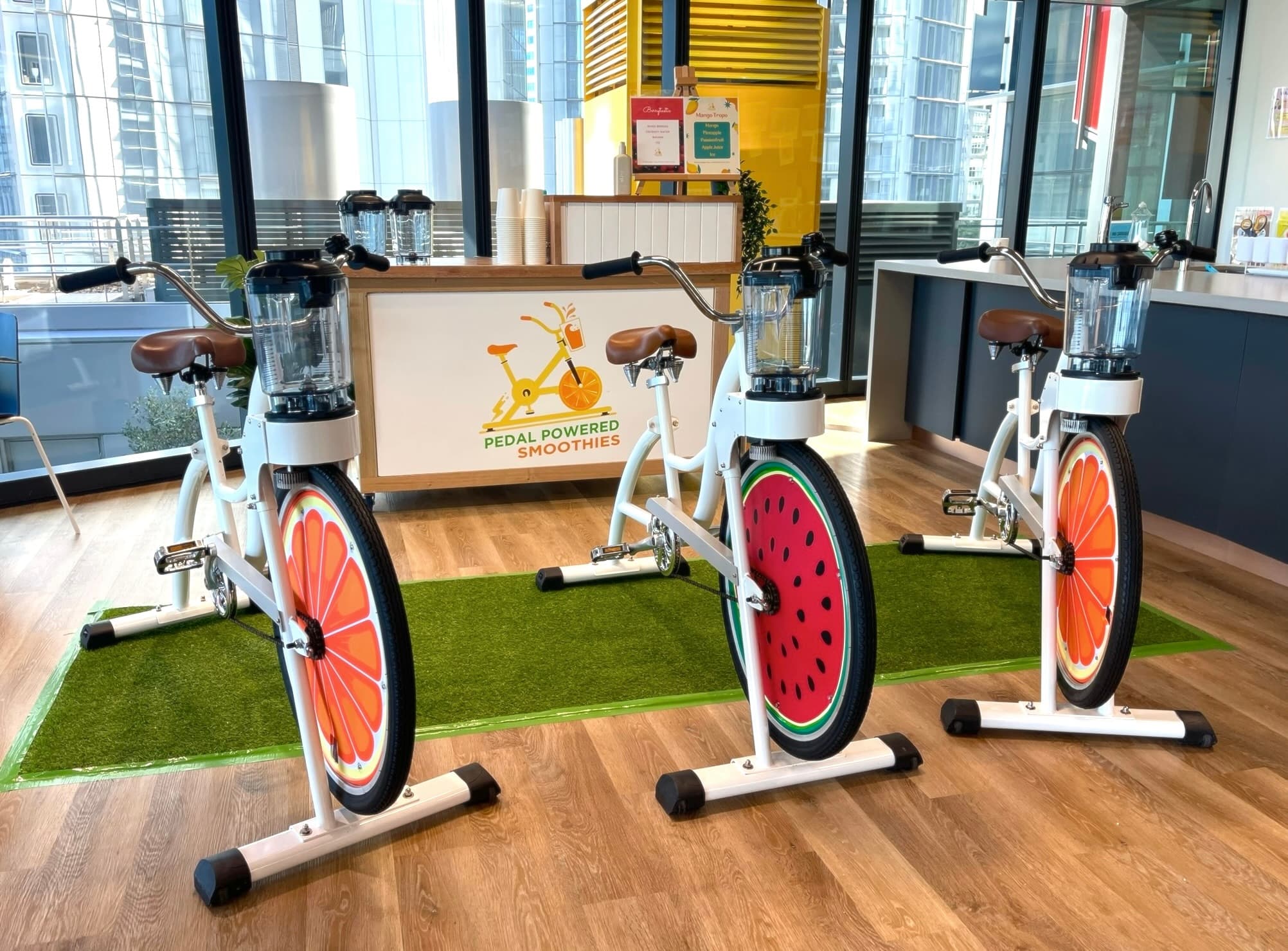 Three stationary bikes with colorful fruit designs on the wheels stand on a green mat indoors. In the background, a “Pedal Powered Smoothies” bar table.