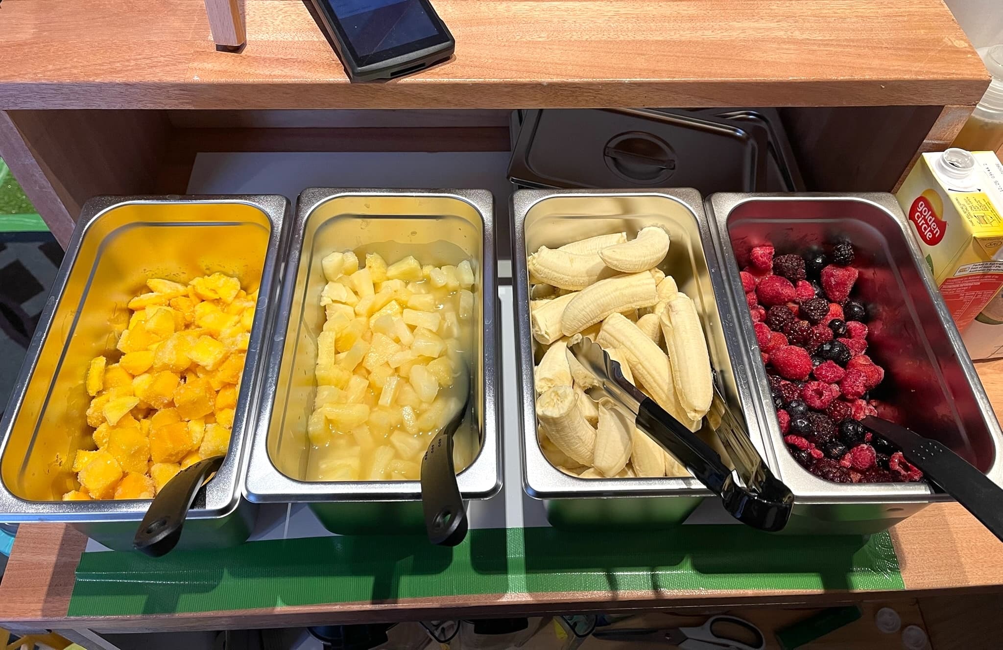 Four stainless steel trays with cut fruits on a counter: mango chunks, pineapple slices, halved bananas, and mixed berries. Tongs rest in each tray.
