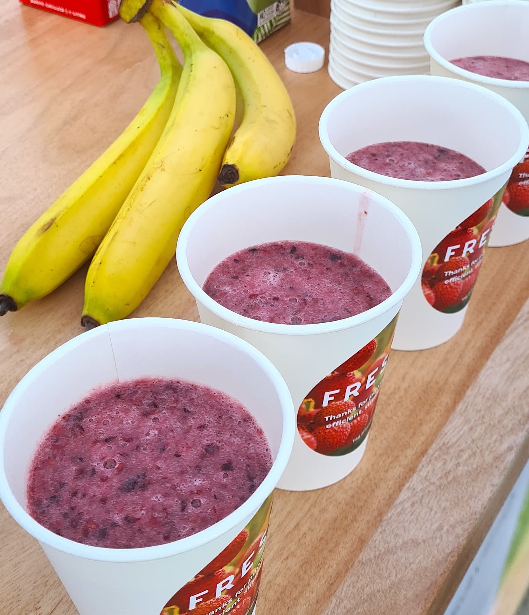 A row of cups of purple smoothies on a wooden table, beside three bananas and juice cartons.