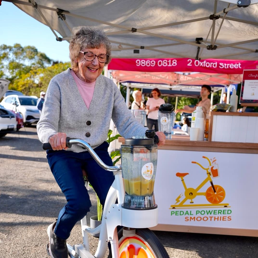 Elderly woman smiles while pedalling a smoothie blender bike at a outdoor market stall.