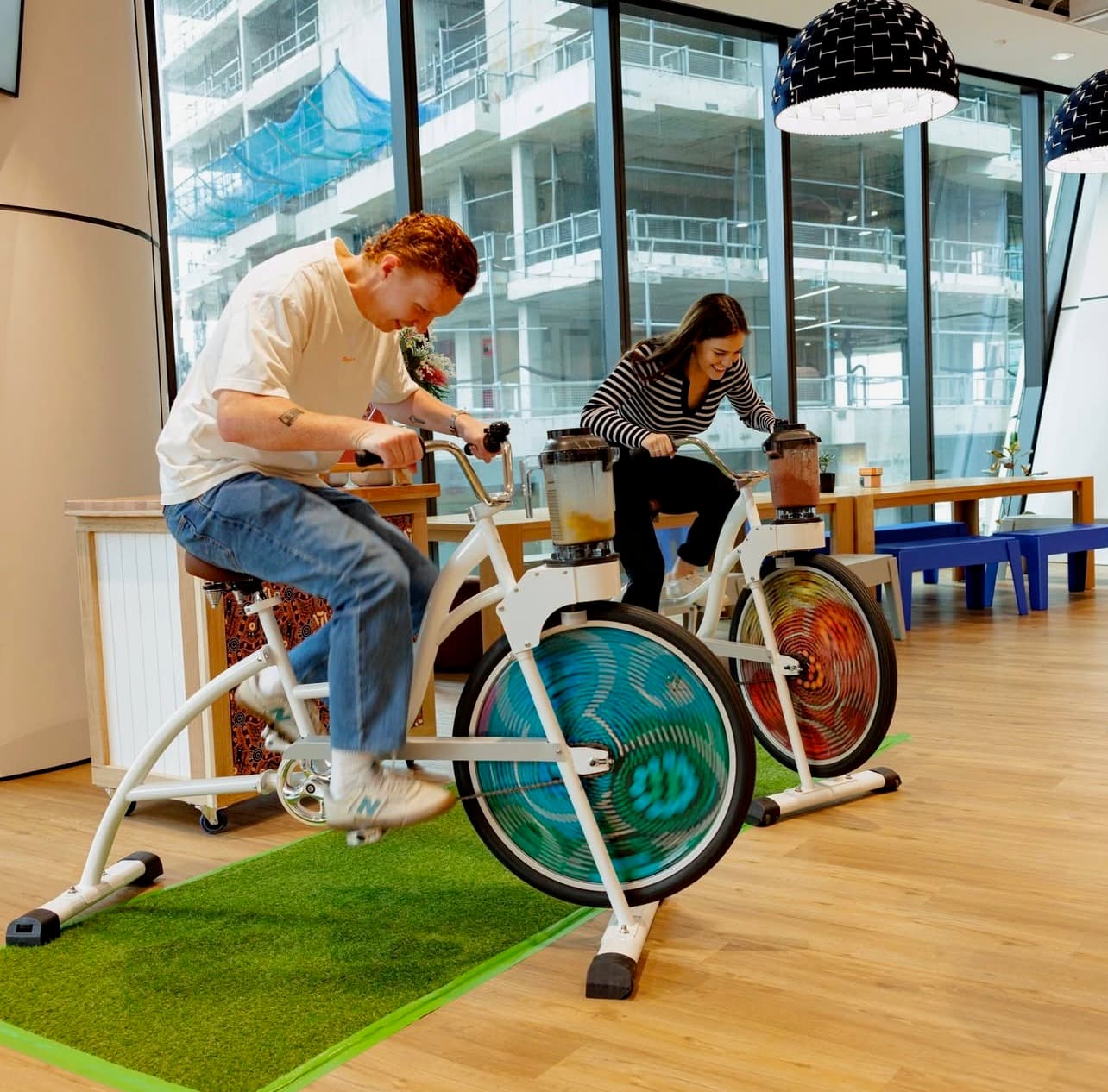 Two people on bikes with colourful wheels, blending smoothies atop the bikes in a modern, glass-walled interior space.