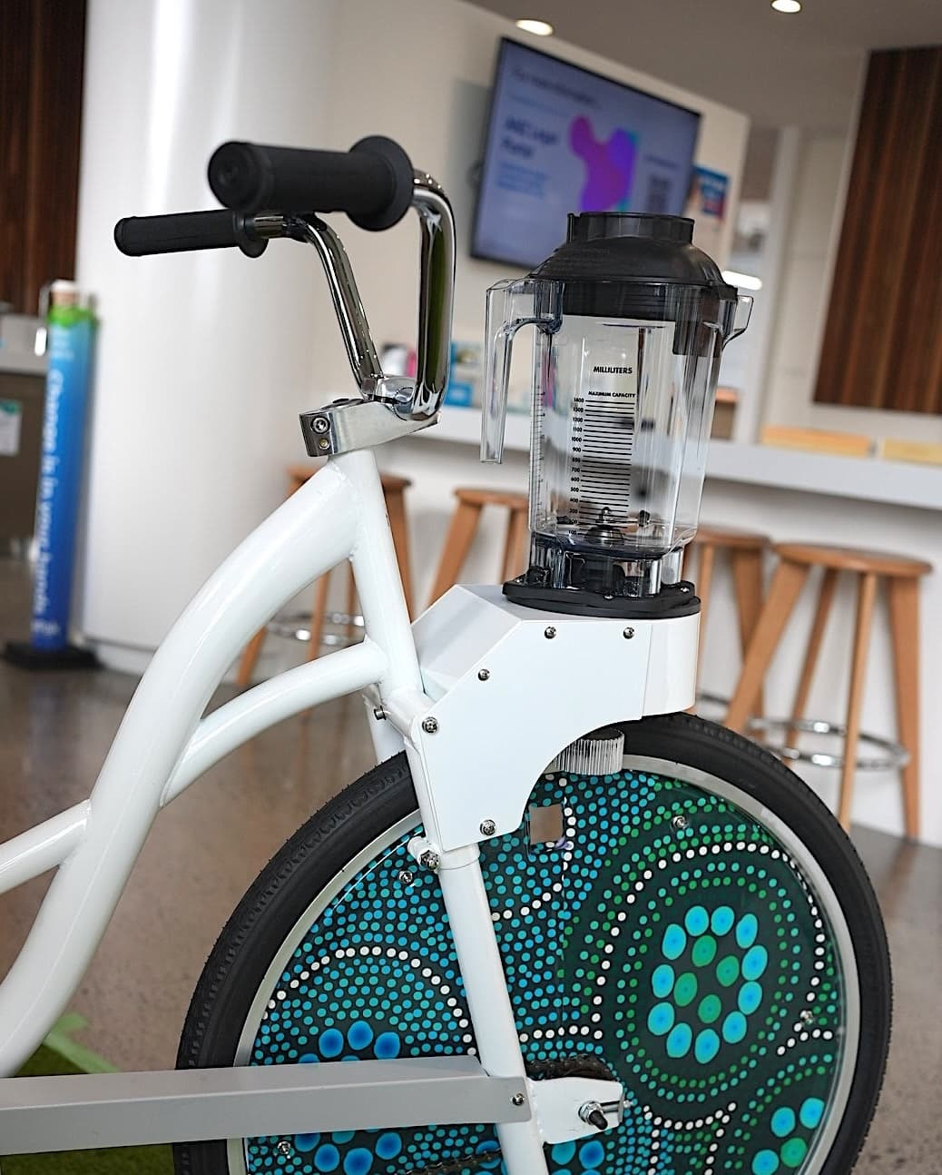 A stationary white bike with a blender attached above the front wheel, in a modern room. The wheel cover is decorated with colorful blue and green indigenous circular patterns.