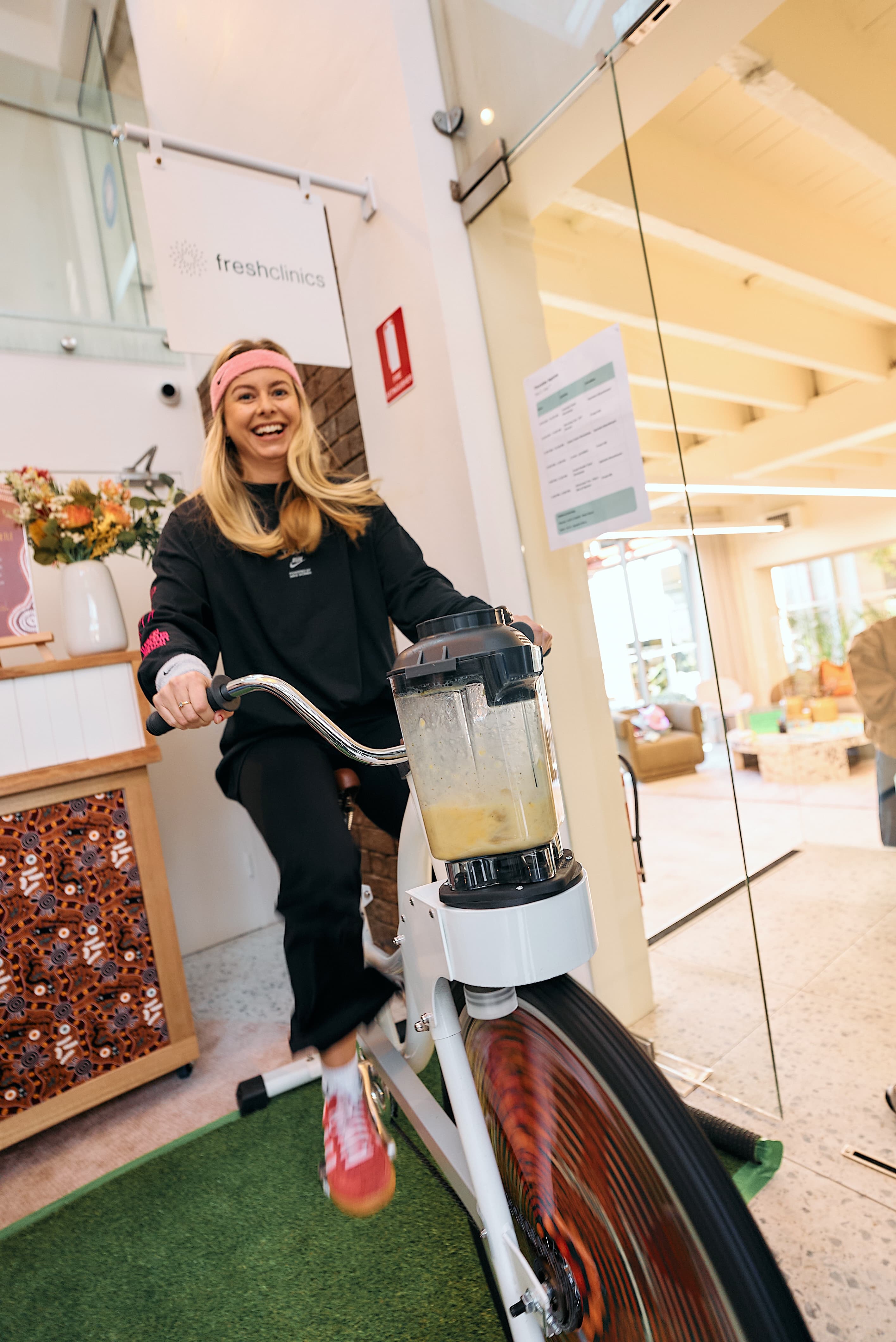 Smiling woman with a headband powering a blender bike. Bright indoor setting with flowers.