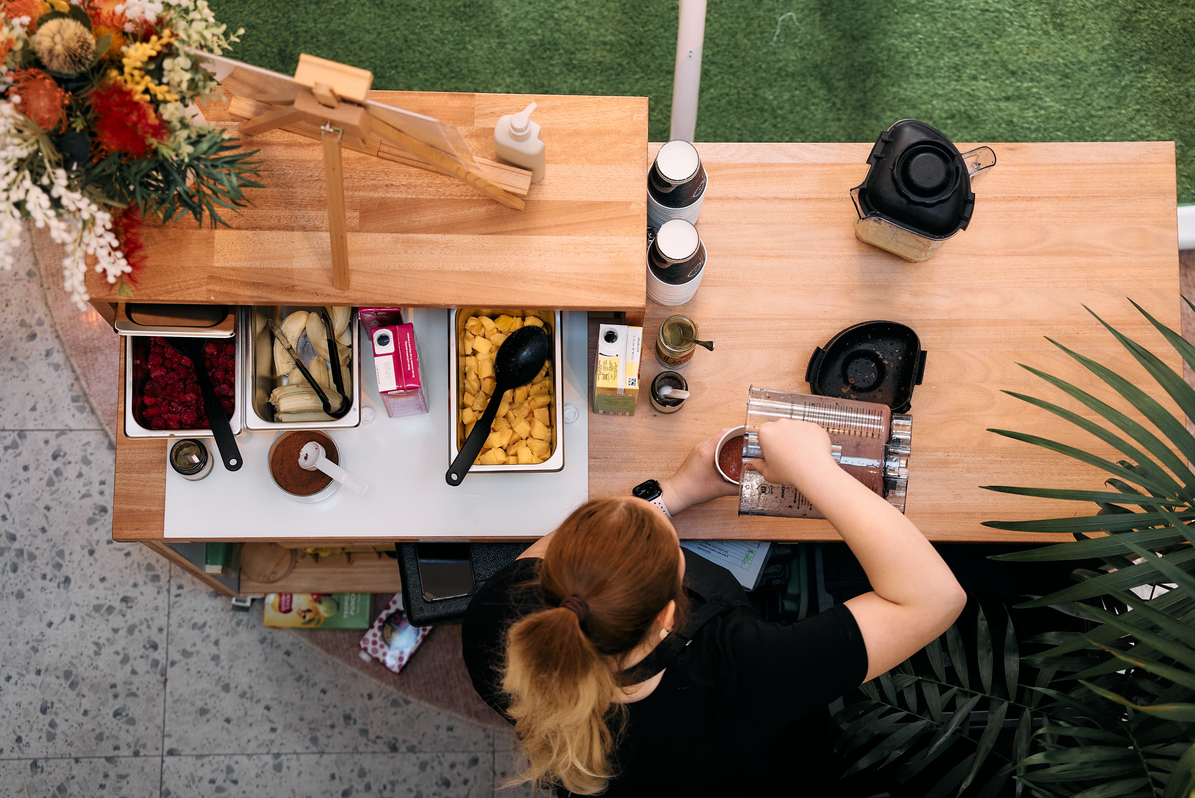 Person prepares a smoothie at a wooden counter with various fruits, cups, and blenders jugs, viewed from above.