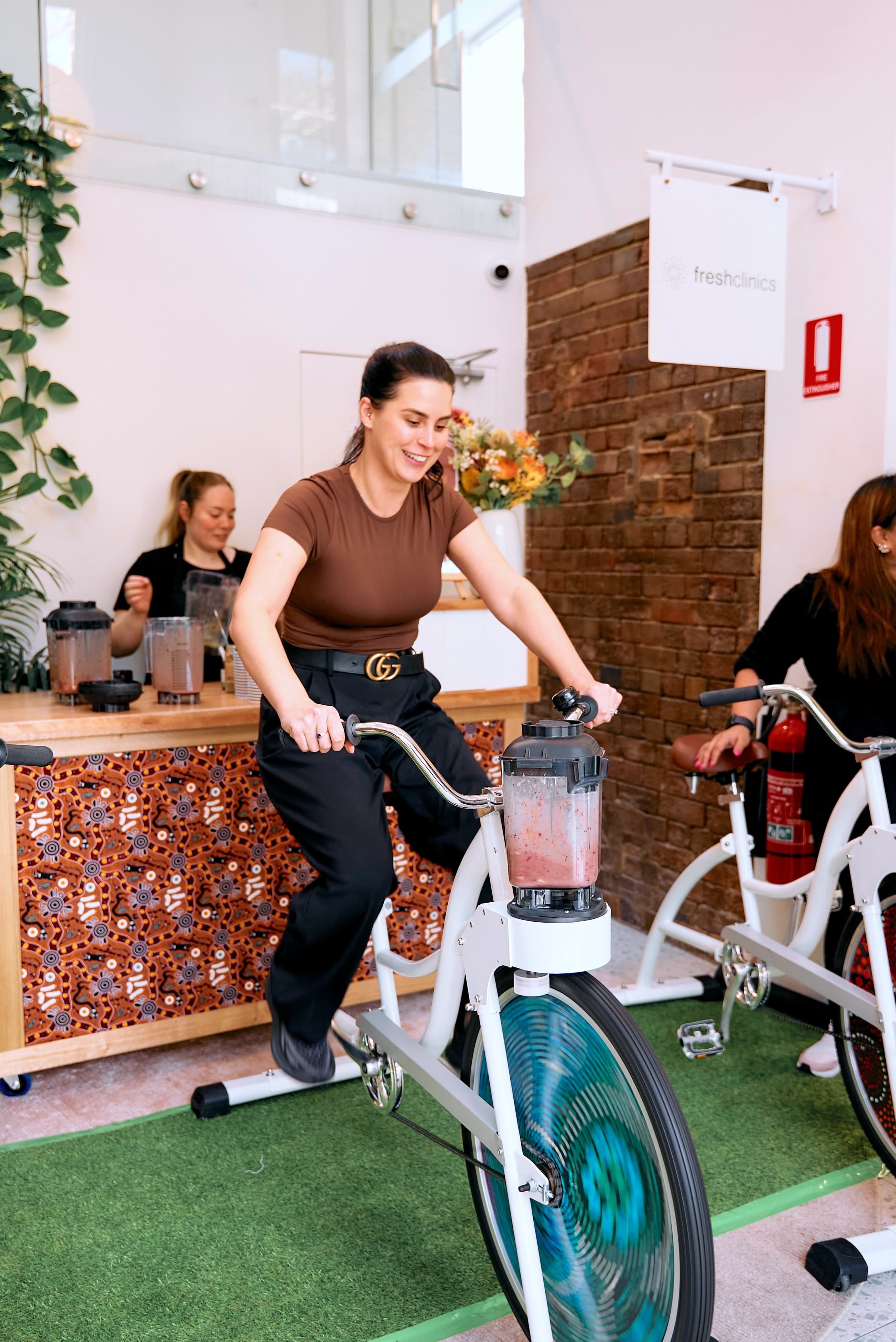 A woman wearing a brown top pedals a bike powering a blender with smoothie ingredients. She smiles, creating an upbeat atmosphere.