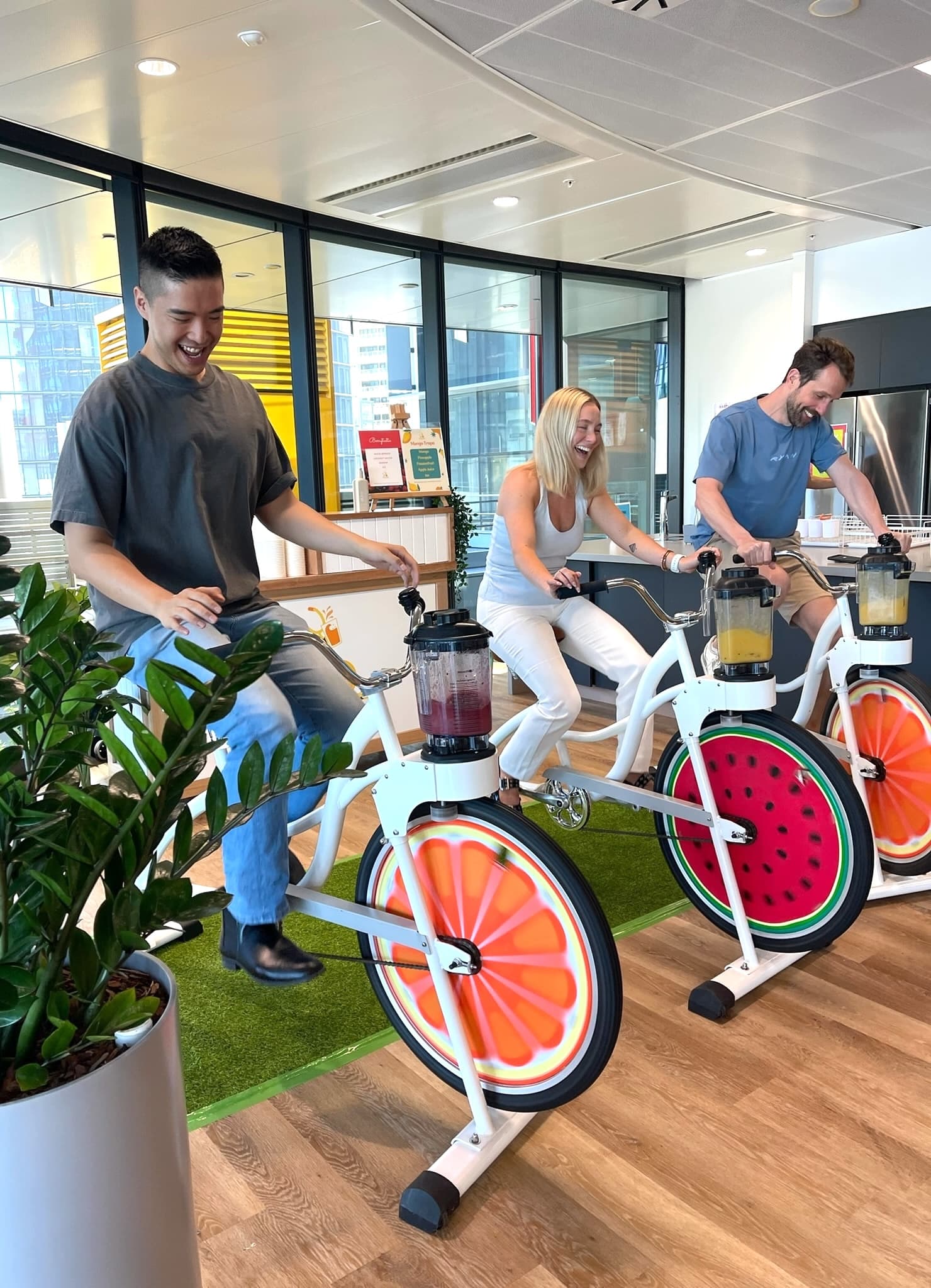 Three colleagues—two men and a woman—are laughing as they sit side-by-side on white blender bikes in a sunlit office kitchen. They are pedalling fast to mix their drinks, with the blender jugs showing vibrant purple and yellow fruit smoothies. The bikes feature the signature orange and watermelon wheel designs.