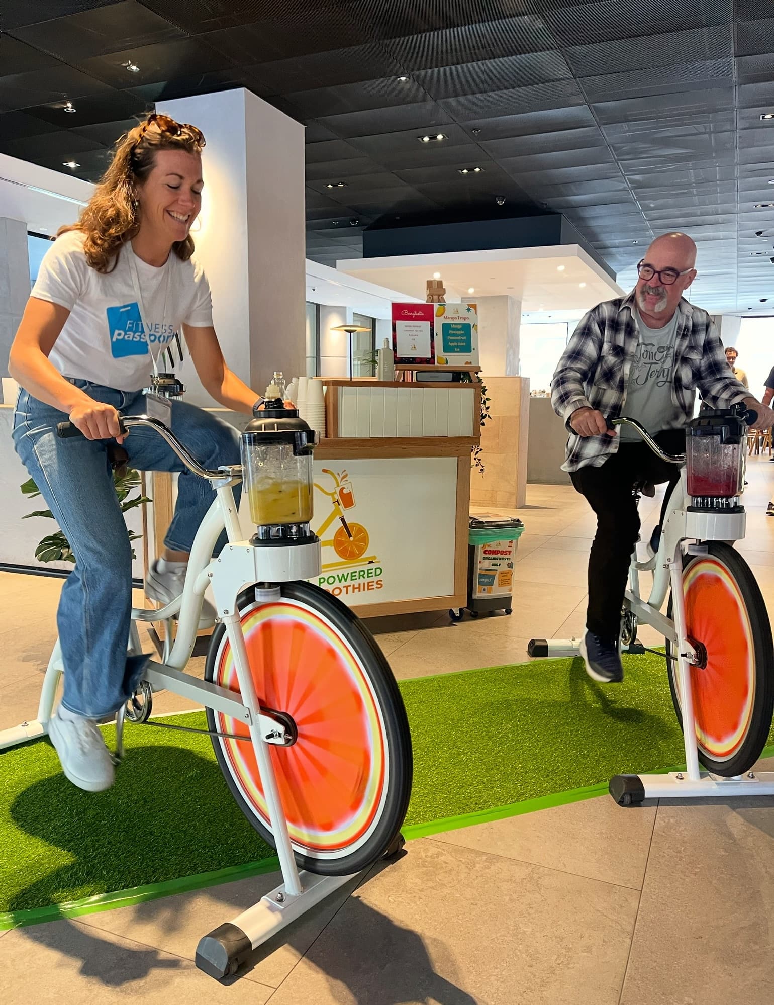 A woman in a "Fitness Passport" t-shirt and a man in a flannel shirt smile while pedalling side-by-side on stationary "Pedal Powered Smoothie" bikes indoors. Each bike has a blender attached to the front handlebars, mixing colorful fruit smoothies as they pedal.