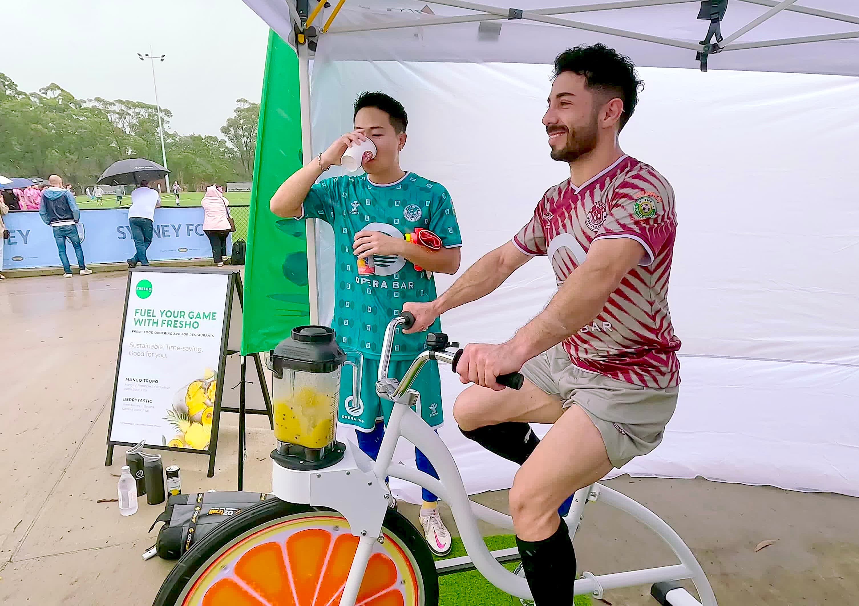 A man in a maroon and white striped jersey pedals a smoothie bike while looking off-camera with a smile. Next to him, another man in a green jersey drinks from a paper cup. They are located at an outdoor sports event with a "Sydney FC" banner in the distance.